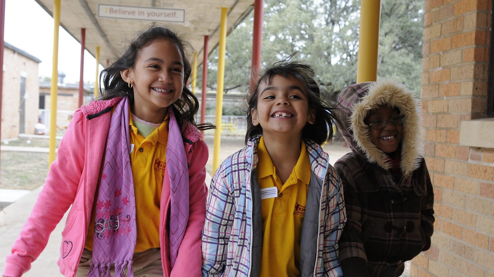 Three elementary school children walk to class smiling in winter coats.