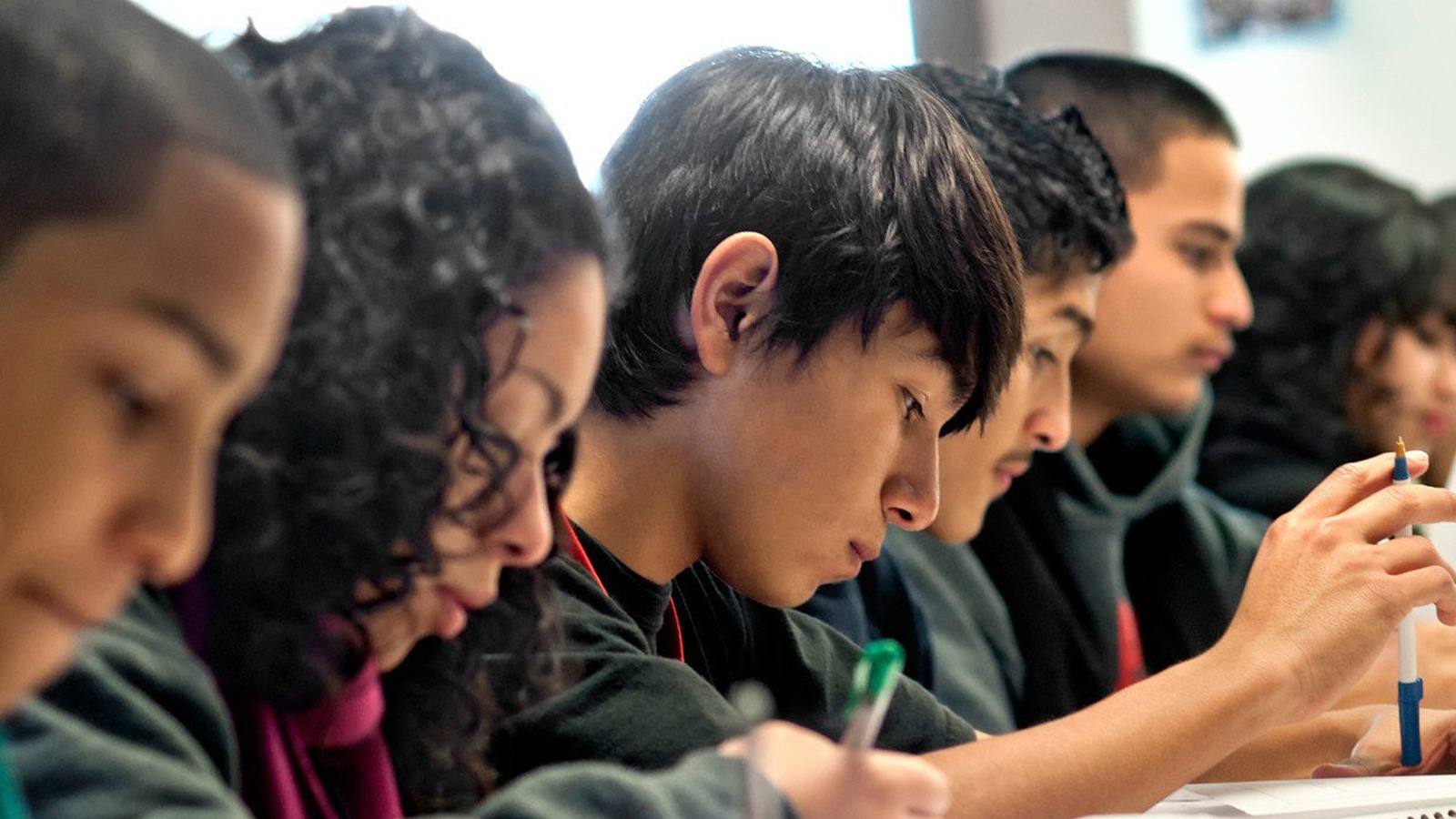 Seven students work at desks next to each other in a row