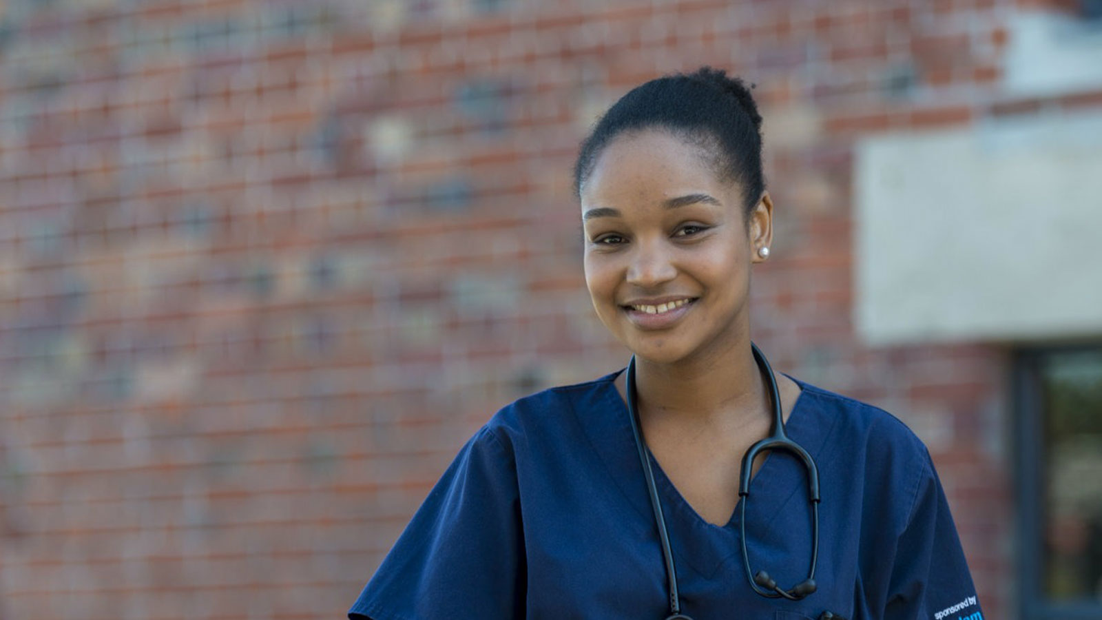 Lindsey Sadie stands outside a healthcare facility wearing doctor's scrubs