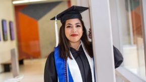 A college graduate stands in a cap and gown by a pillar.