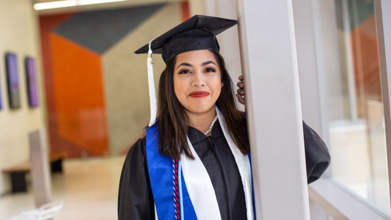 A college graduate stands in a cap and gown by a pillar.