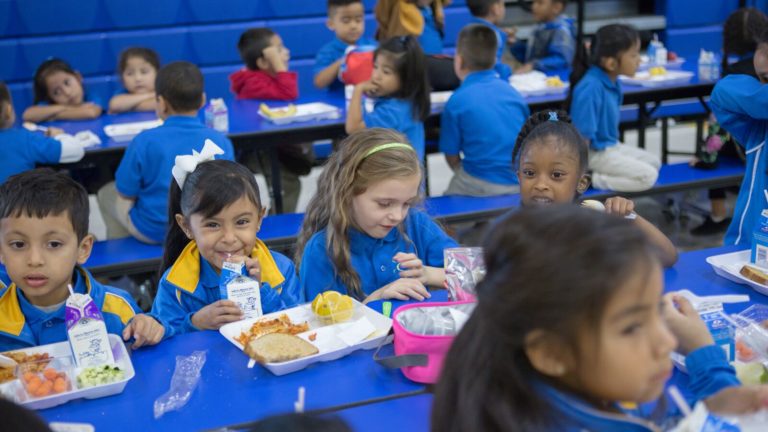 Elementary schools students eat lunch at a table in the school cafeteria.