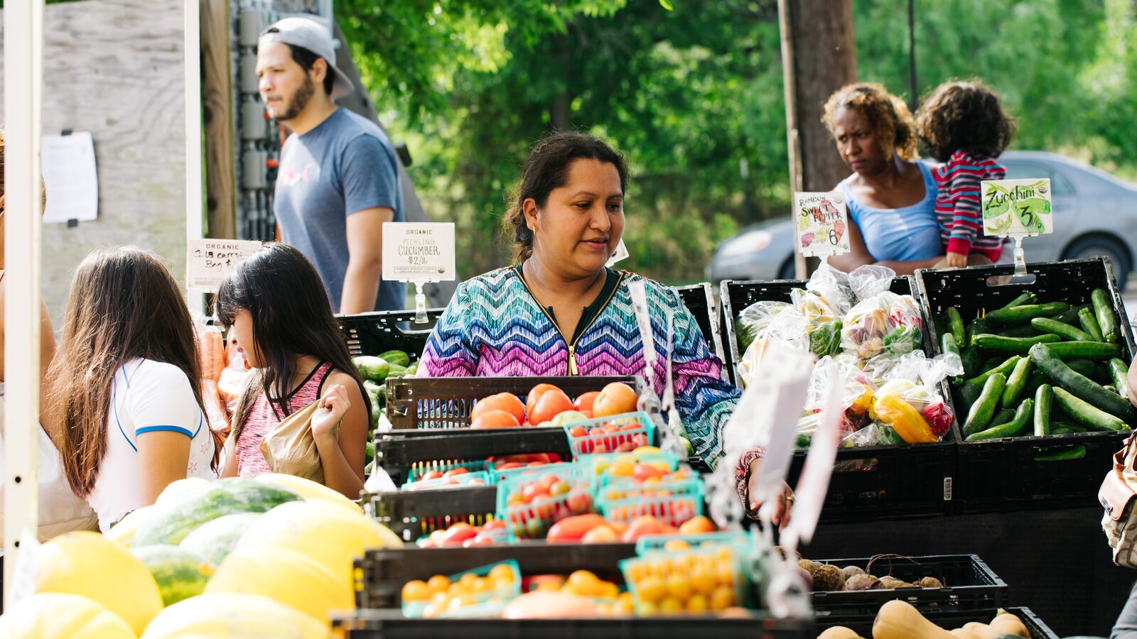 A woman at a farmer's market looks at affordable fruit and vegetables next to families