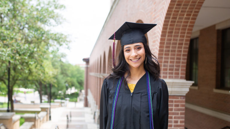A college grad walks on her college campus in cap and gown.