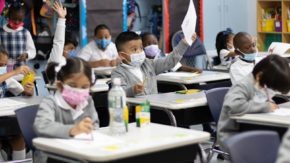 medium shot of classroom full of elementary students working at their desks wearing masks. One student in the center is raising a sheet of paper in the air while students around him continue working