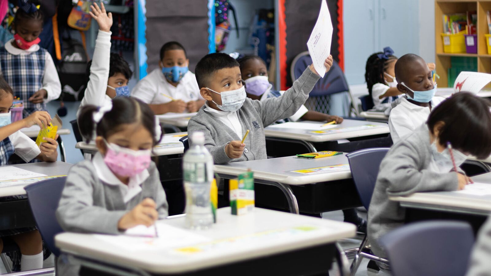 medium shot of classroom full of elementary students working at their desks wearing masks. One student in the center is raising a sheet of paper in the air while students around him continue working
