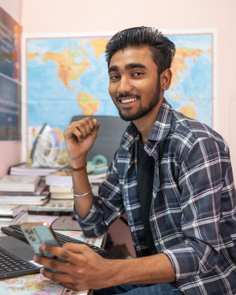 Photo of student, Abhishek, sitting at a desk with his phone, smiling direct to camera.