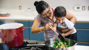 A woman and a child in a kitchen standing over a stove and stainless steel pan stirring sauteed vegetables with a silicon spatula.