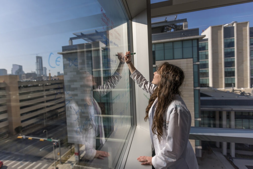 Student, woman in white lab coat writing on a window with marker overlooking city. 