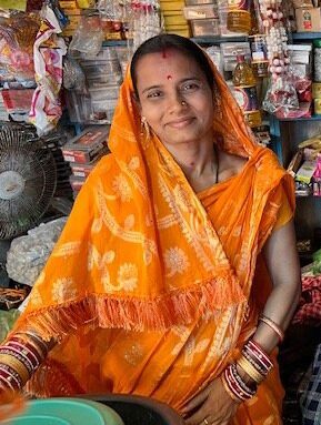 Nano entrepreneur Ambika sits smiling in her small shop. Waring orange sari and scarf surrounded by merchandise.