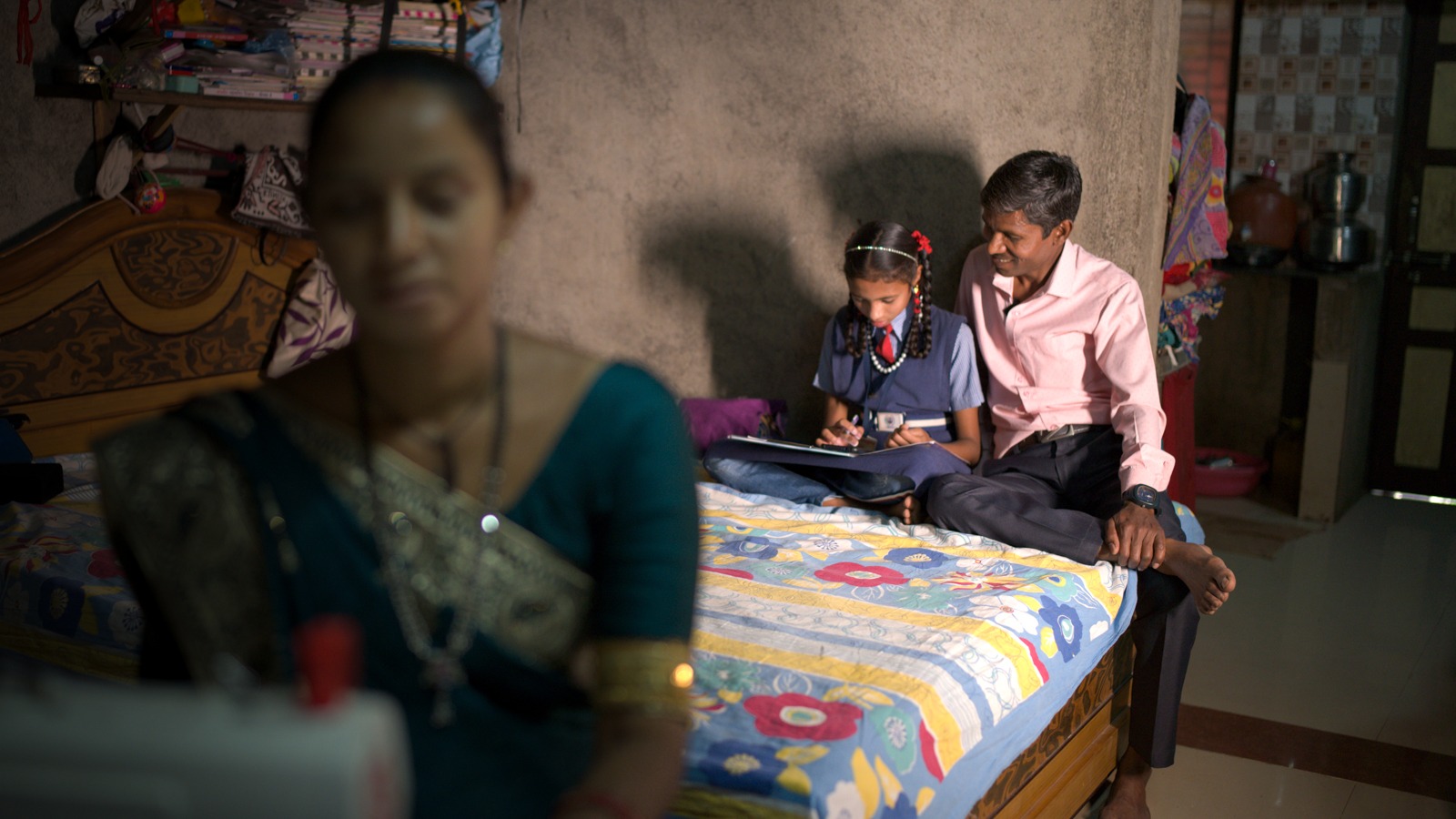 Young girl in a school uniform works on homework on a tablet in her family home with her parents.
