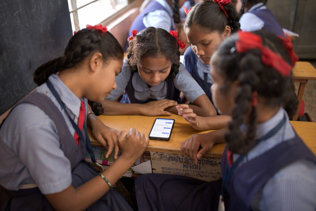 Group of young students, girls, in school uniforms, light blue and grey with red bows, gather around a smartphone reviewing schoolwork.