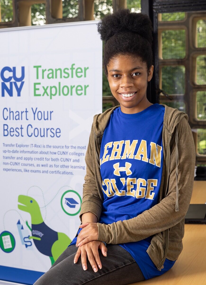 Photo of Yesenia, a CUNY student, sitting and smiling direct to camera in front of a standing banner featuring the Transfer Explorer tool.