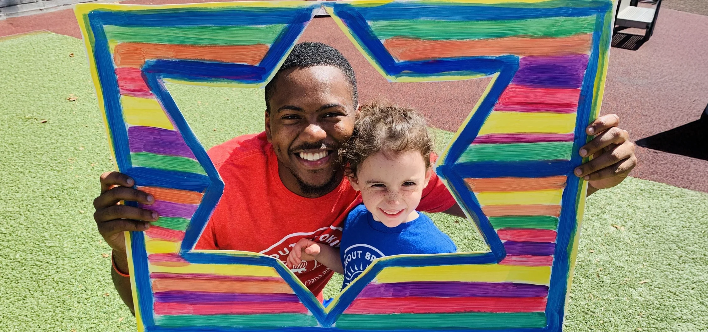 A man and child smile while holding up a colorful sign on a playground.