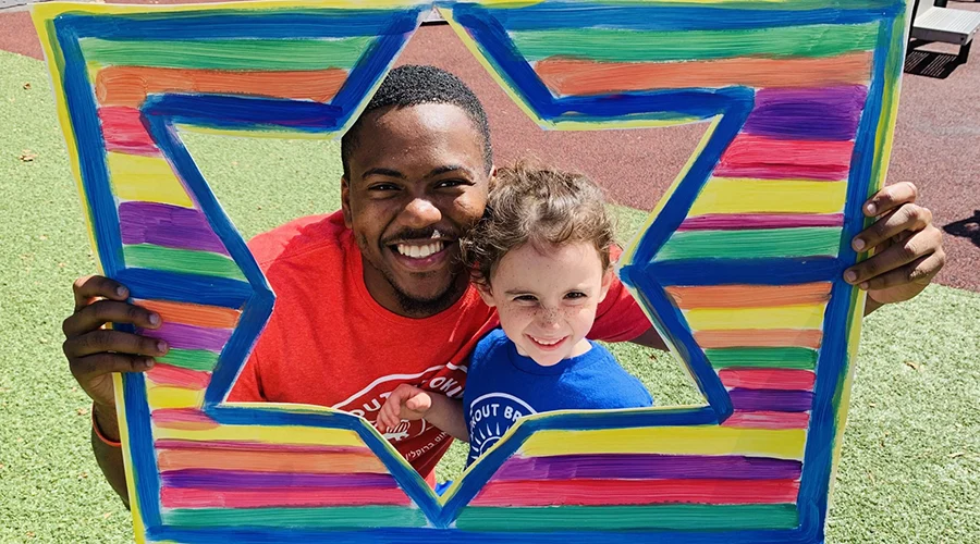 A man and child smile while holding up a colorful sign on a playground.