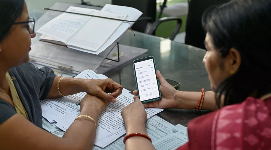 Two women sit at a desk, looking at a phone screen together.