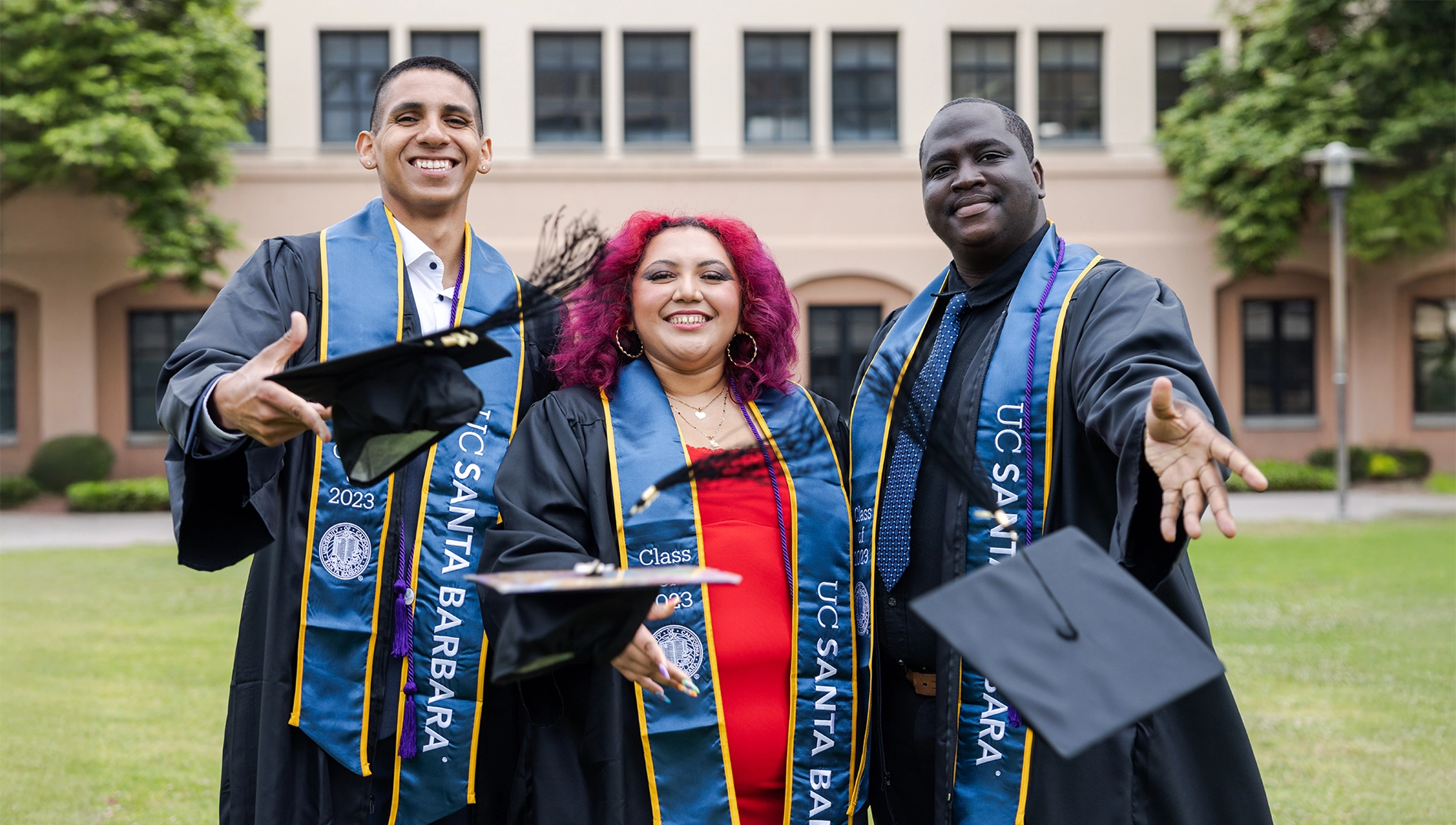 Three students in graduation caps and gowns, throw their caps toward the camera.