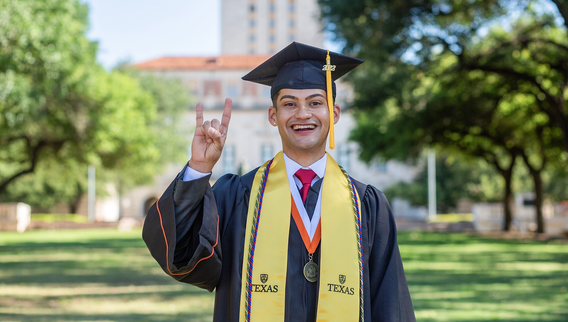 A University of Texas at Austin graduate stands in front of the tower in his cap and gown, giving the hook 'em horns sign.