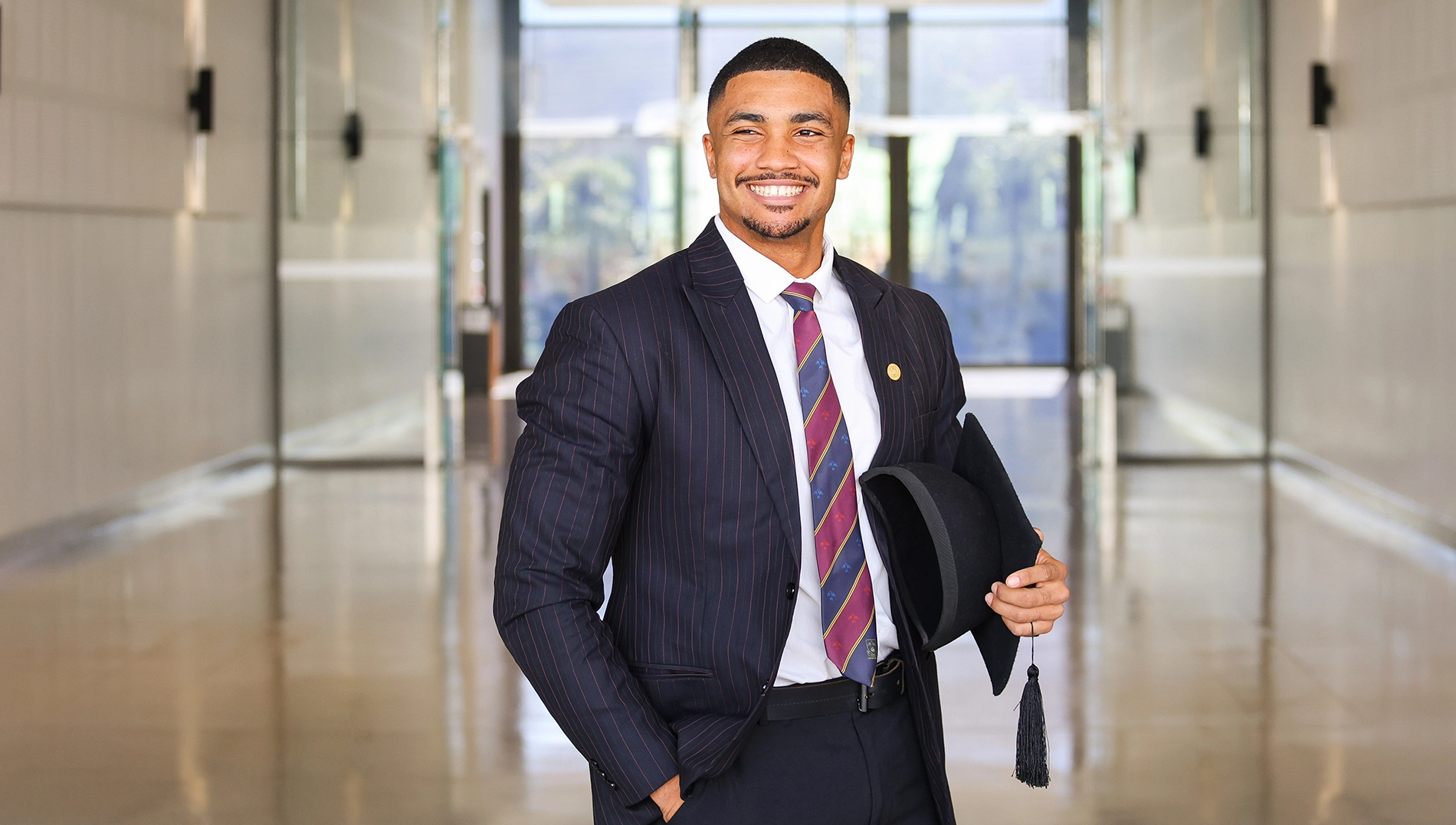 A man stands in an office in suit and tie, holding his graduation cap.