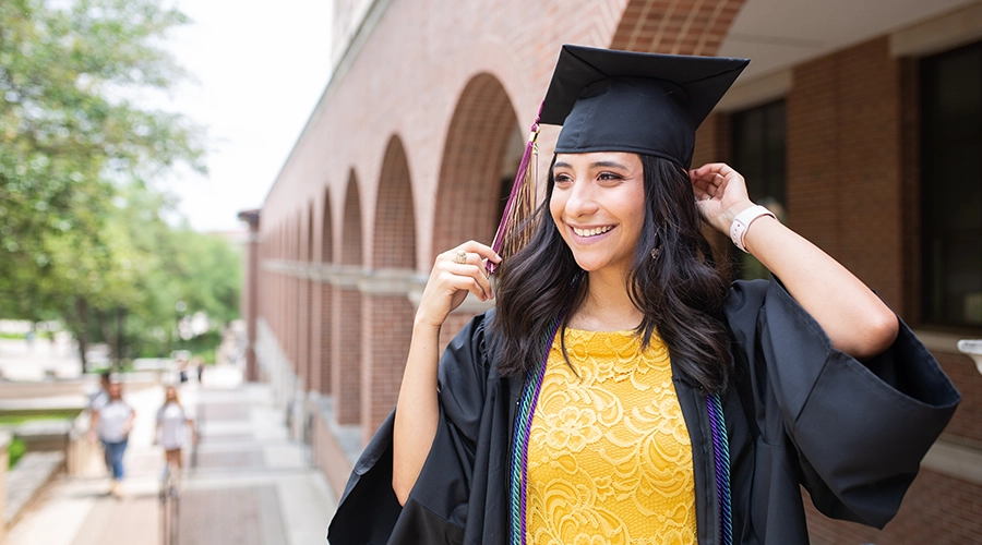 A graduate stands on campus, wearing a cap and gown.