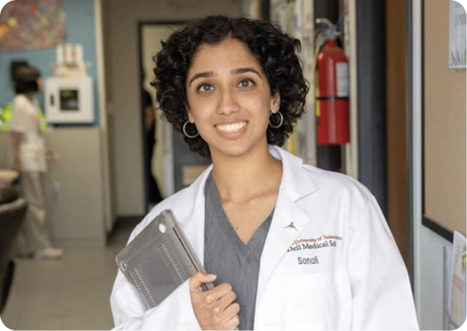 A medical student in scrubs and a white coat holding a laptop in a hospital hallway.