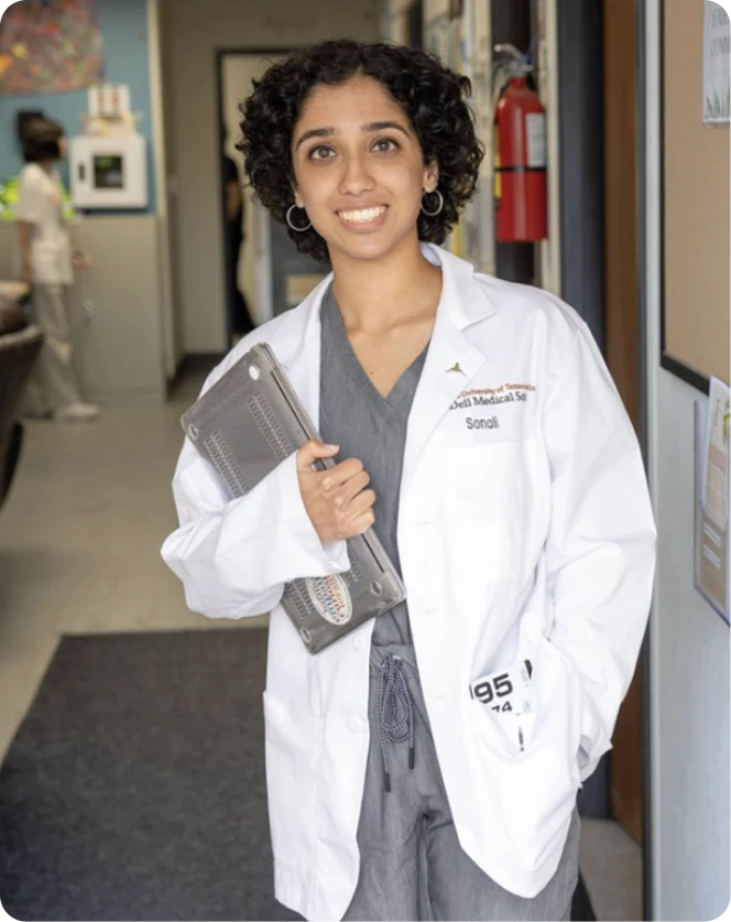 A medical student in scrubs and a white coat holding a laptop in a hospital hallway.