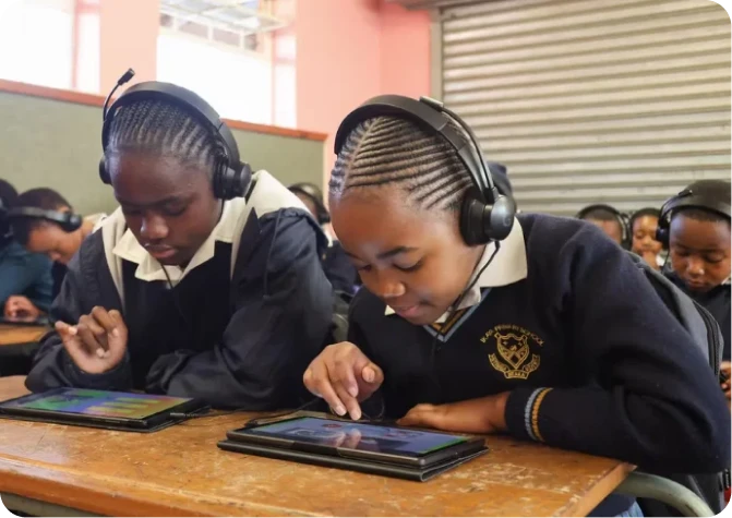 Two students wearing headphones use tablets together in a classroom.