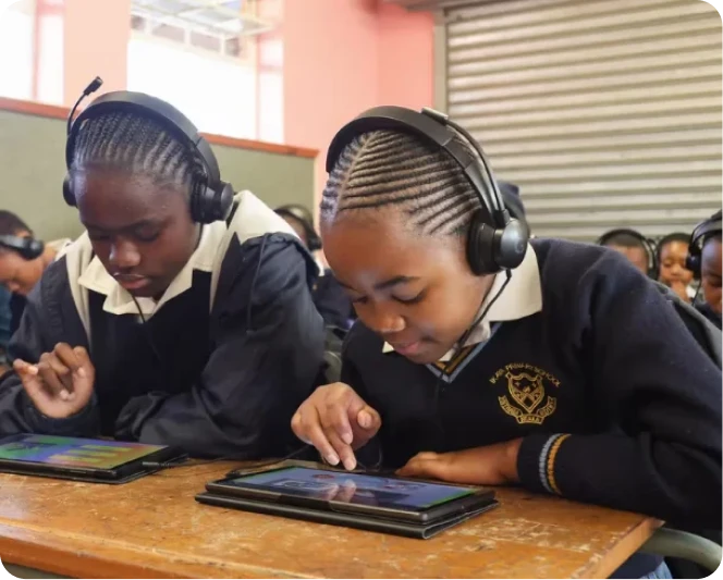Two students wearing headphones use tablets together in a classroom.