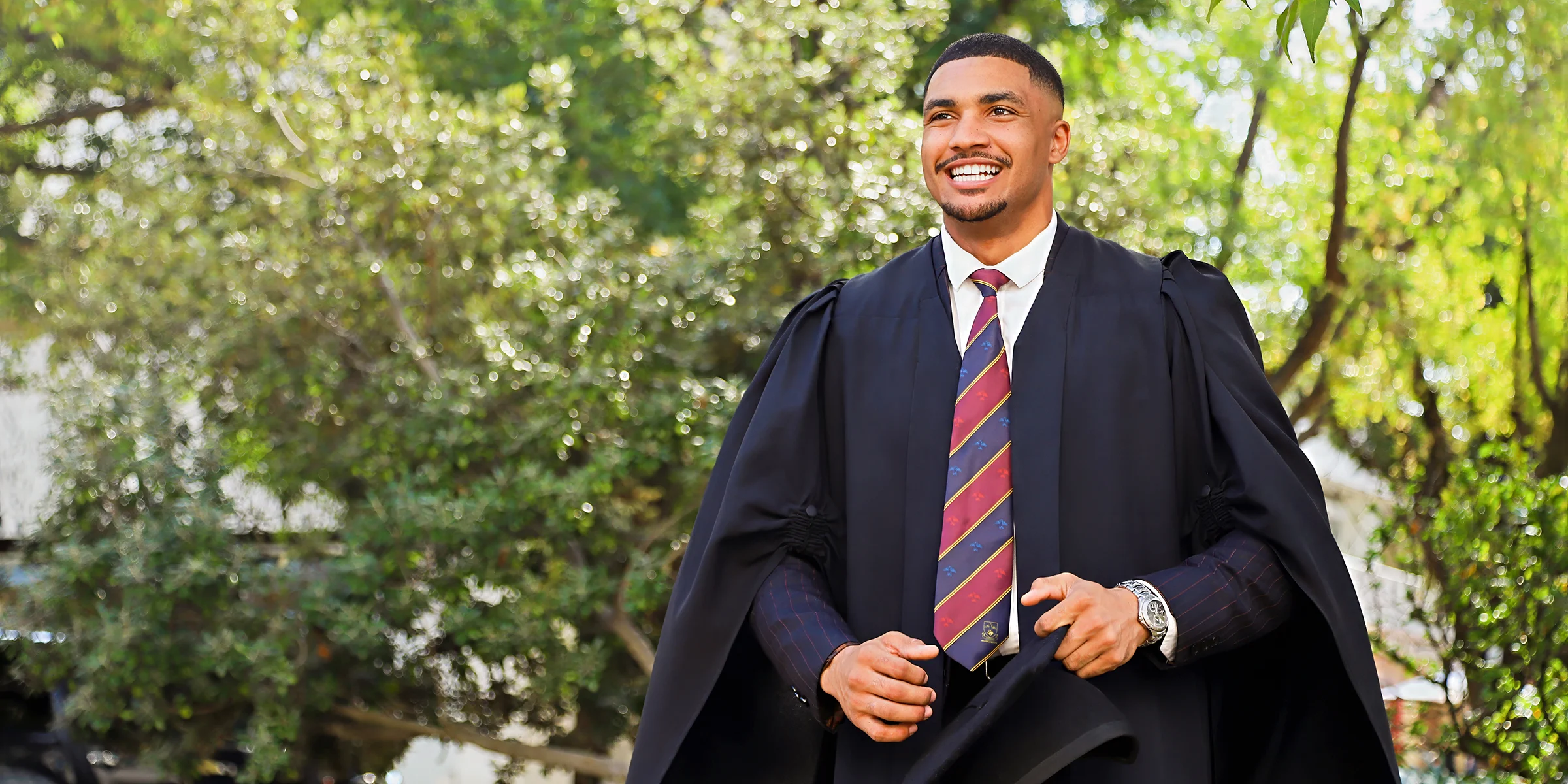 A Dell Young Leader graduate standing in front of greenery while wearing his graduation gown and holding his cap in hand.